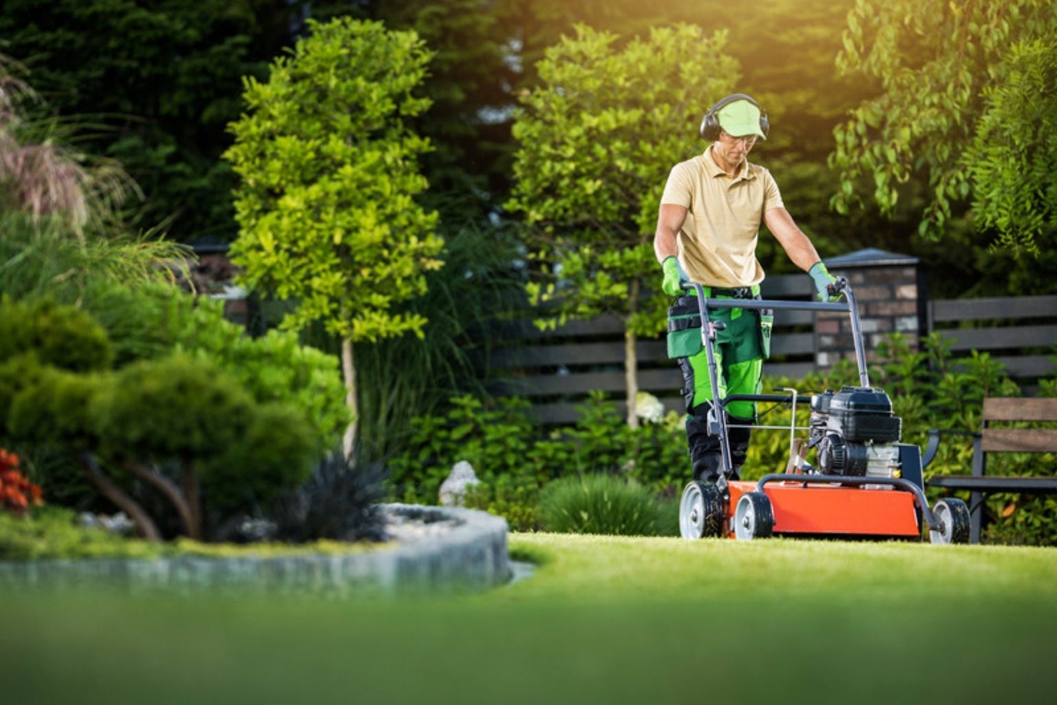Gartner med høreværn slår græs med en rød plæneklipper i en velplejet have